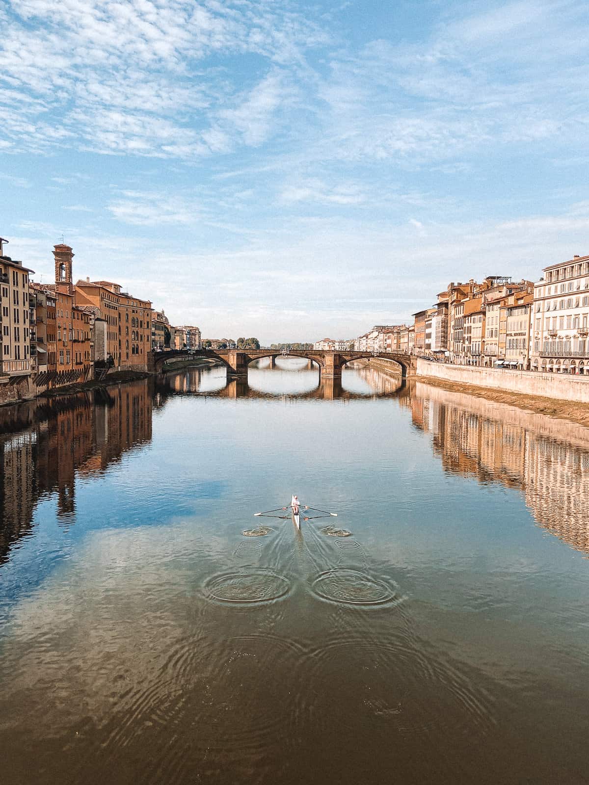 A serene view of Florence's Arno River, with a lone rower in the center creating ripples on the water's surface, framed by historic buildings and the iconic Ponte Vecchio in the distance under a clear blue sky.