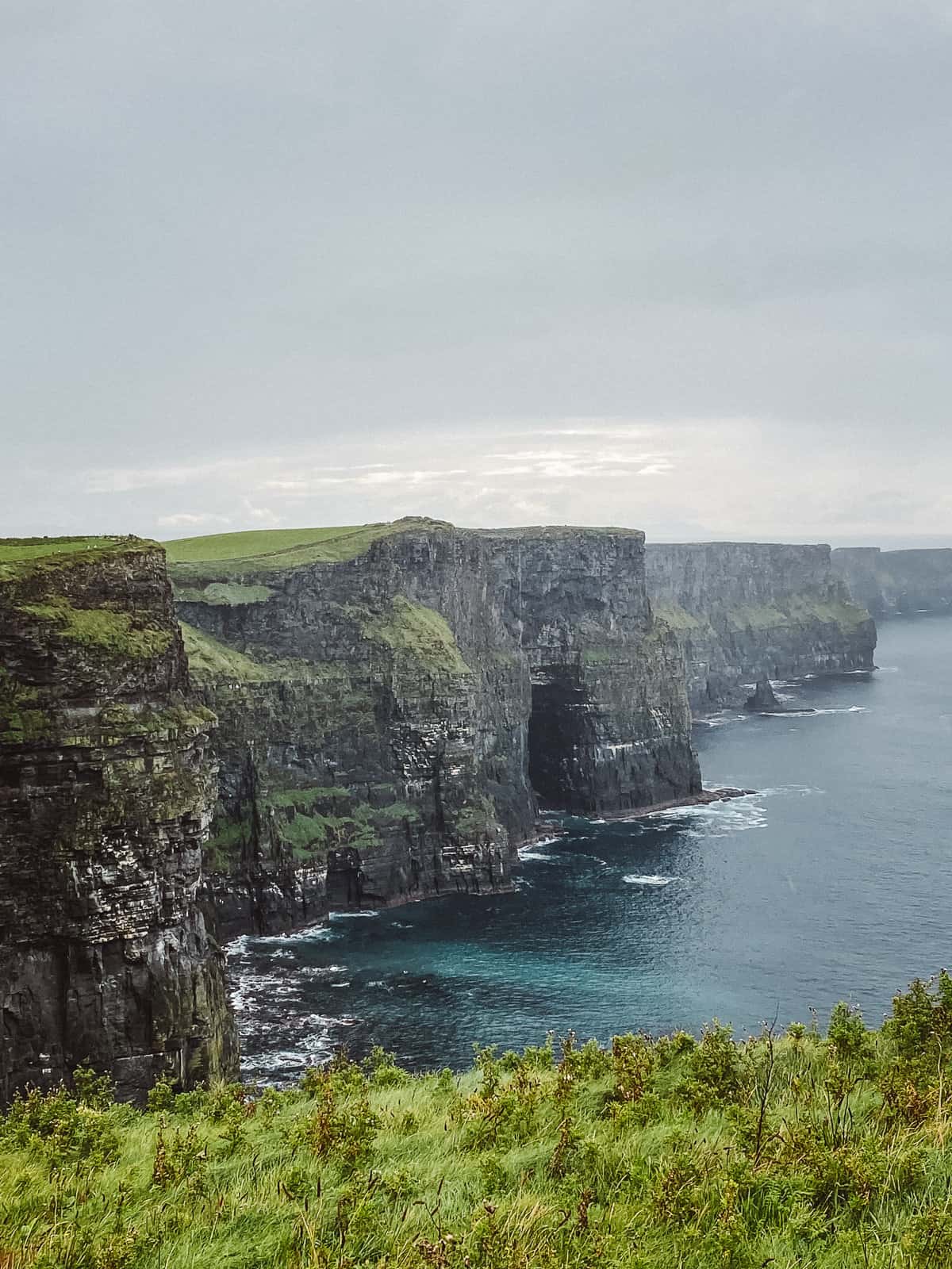 The lush green landscape of Ireland's Cliffs of Moher, with the sea cliffs dropping dramatically into the Atlantic Ocean, under a moody sky that highlights the natural beauty and ruggedness of the Irish coastline.