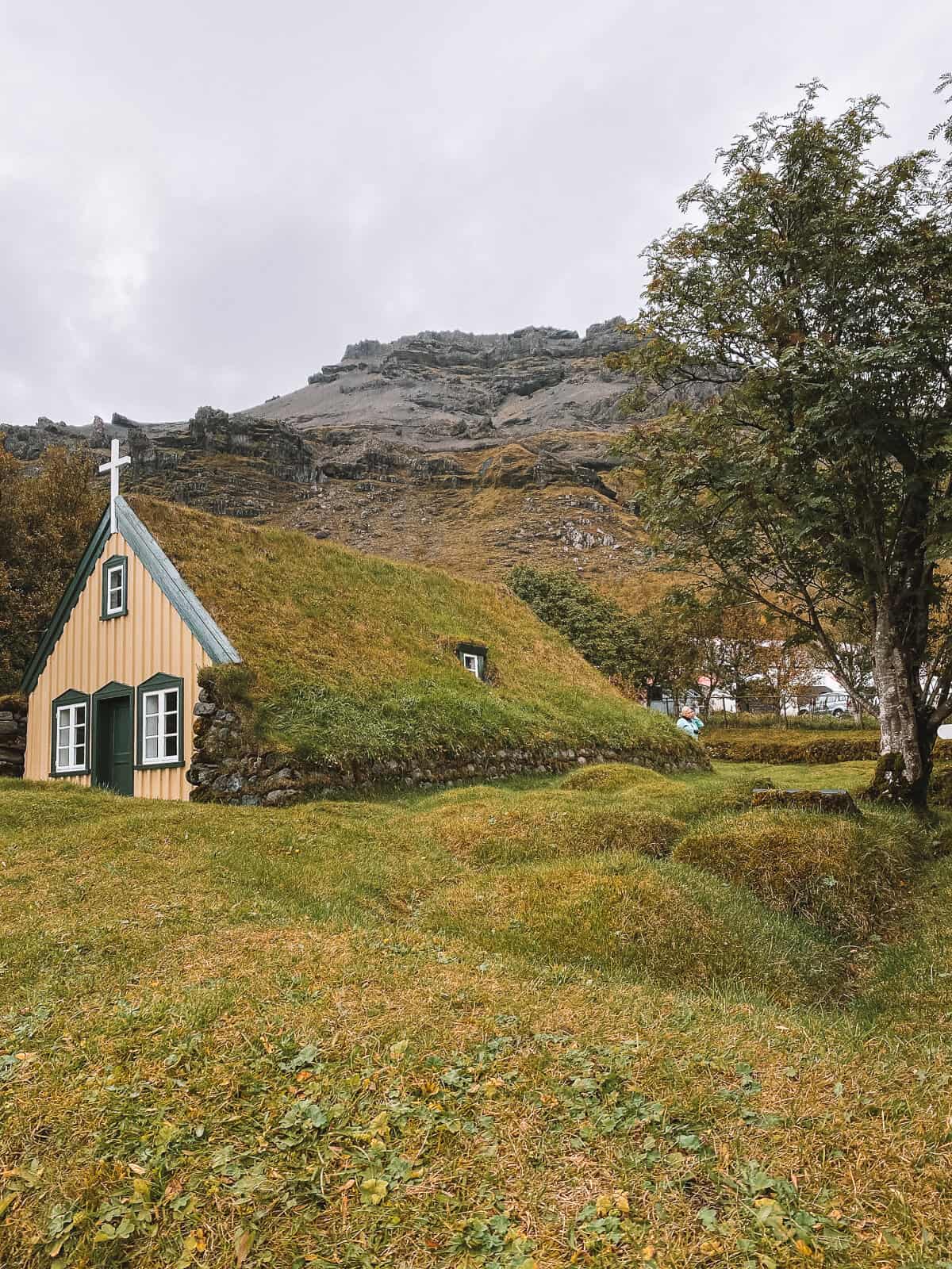 A traditional Icelandic turf church with a green roof, nestled against a backdrop of rugged mountains, portraying the quaint charm and symbiosis with nature characteristic of Icelandic architecture.