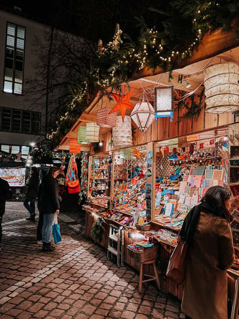 A Christmas market booth under a large tree strung with lights, offering colorful paper lanterns and assorted crafts, with shoppers browsing in the evening light.