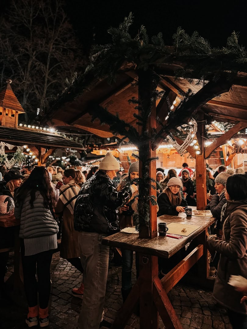 Patrons gathered around a wooden stall at a Christmas market, enjoying drinks and conversation under a roof adorned with festive greenery and lights.