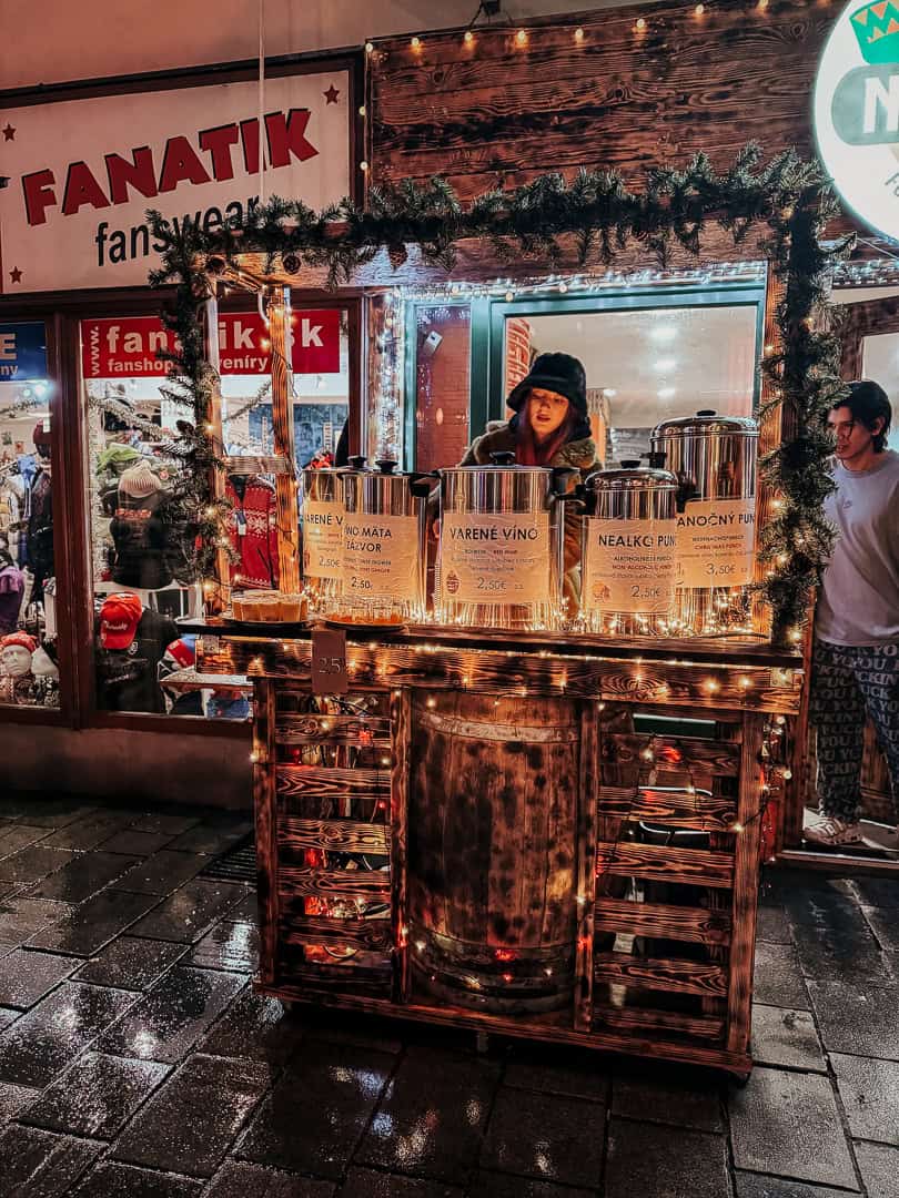 A cozy drink stall at a night market, warmly illuminated, selling mulled wine and punch with prices displayed in euros, as a woman in a winter hat serves customers.