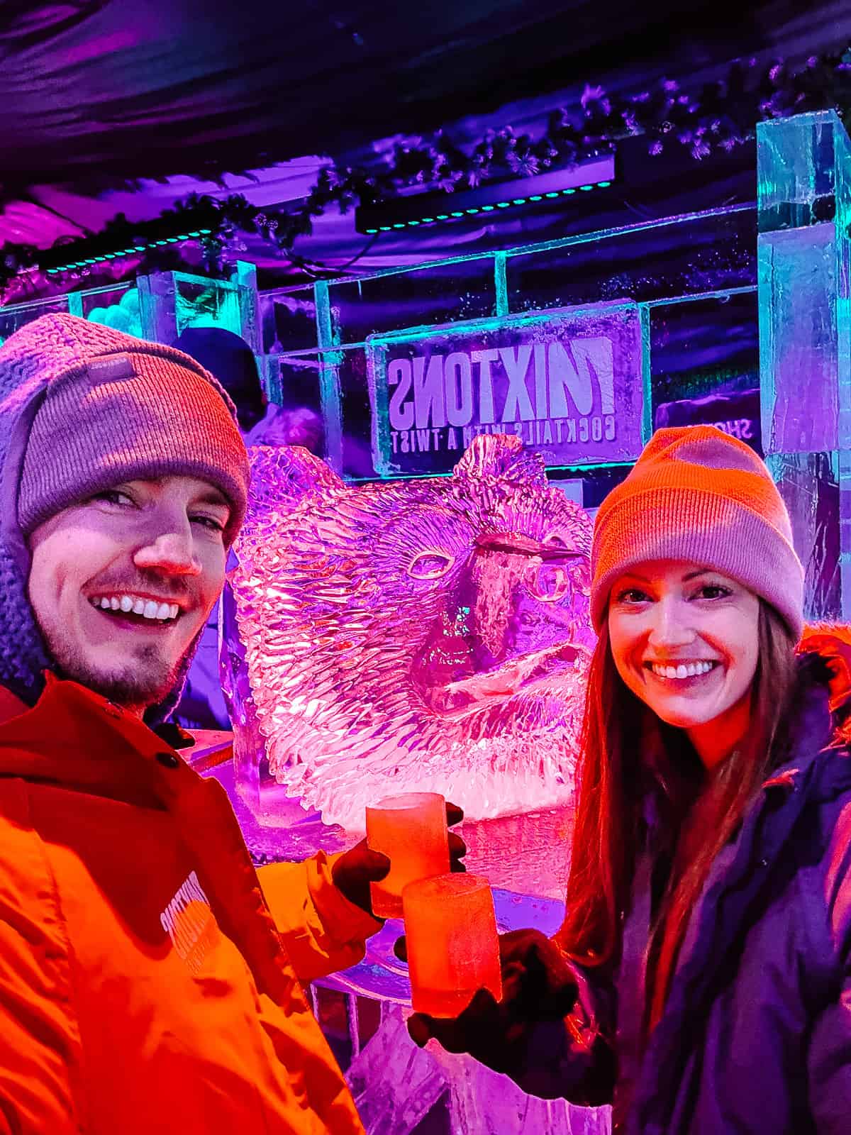A man and woman share a toast with drinks in ice glasses, surrounded by the glowing purple ambiance of an ice bar