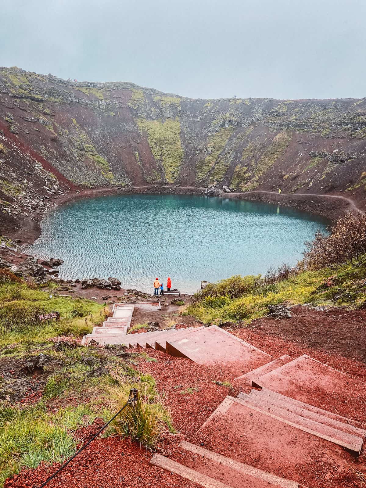 Descending steps leading to a serene volcanic crater lake in Iceland with lush green moss-covered slopes under a misty sky, with two hikers in bright jackets standing by the water's edge, enhancing the tranquil yet vibrant scene.