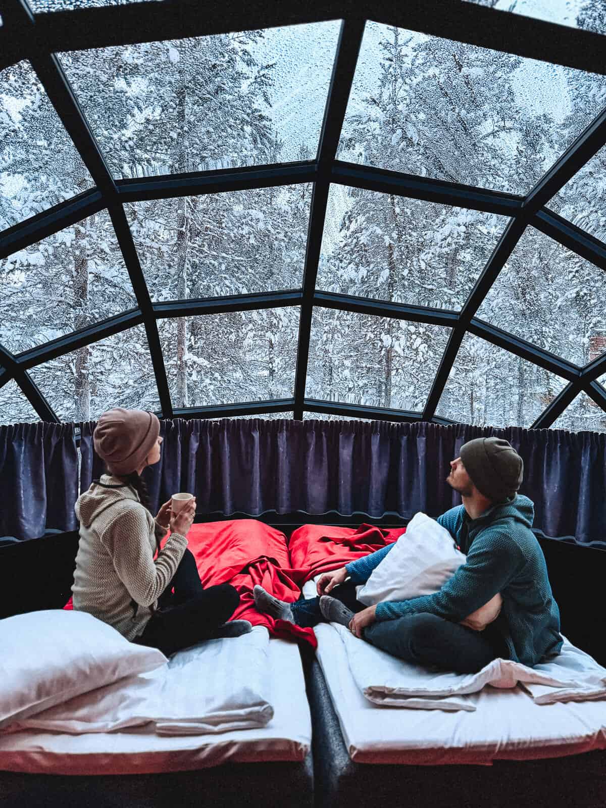 Two people relax inside a cozy igloo with a transparent dome, sipping warm drinks and looking out at a snowy forest landscape