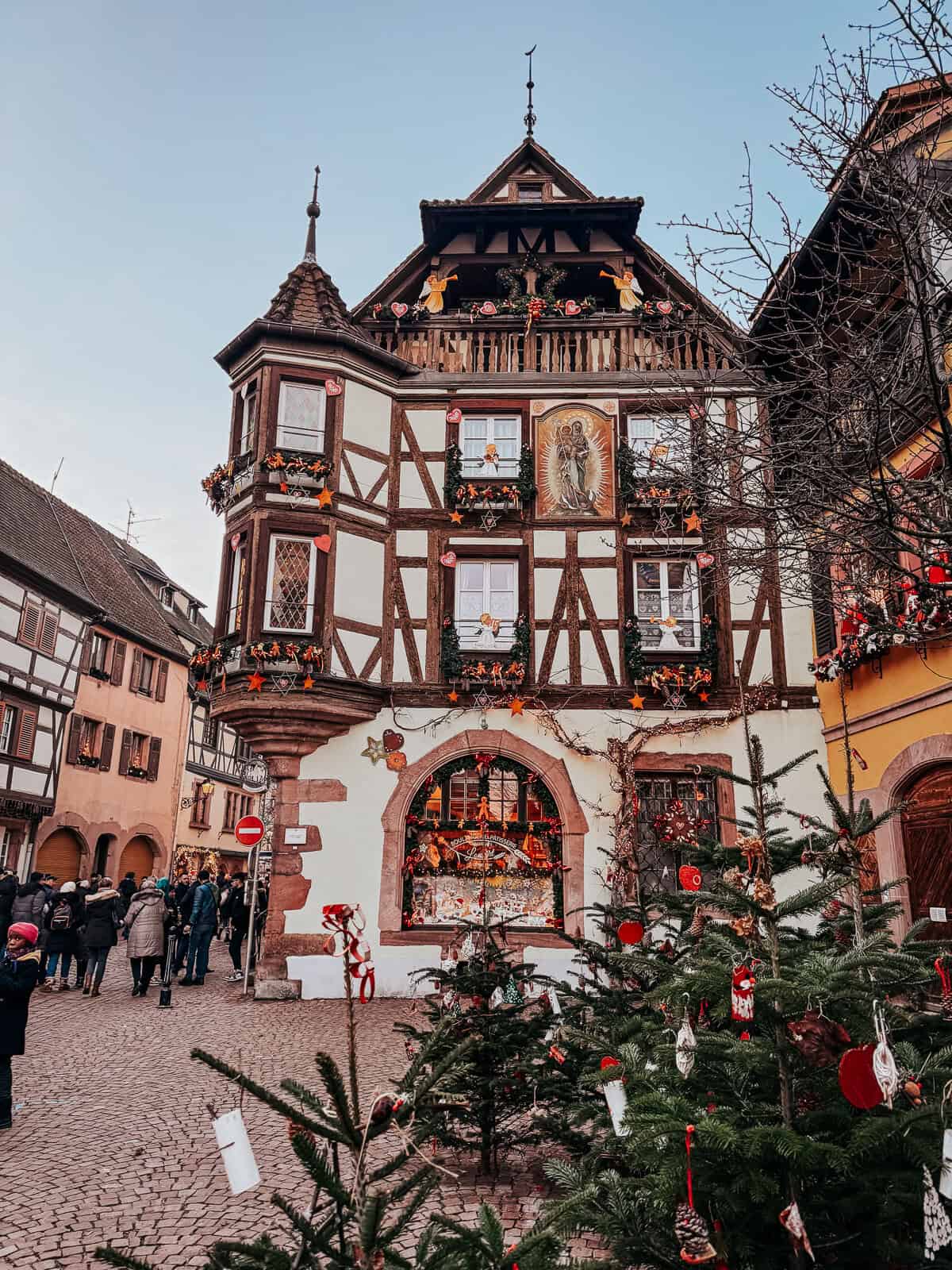 A beautifully decorated corner in Kaysersberg with a Christmas tree and festive ornaments, set against a historical half-timbered house with murals and wreaths.