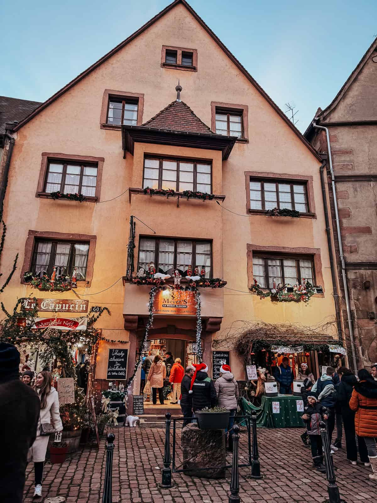 Visitors gather in front of a festive peach-colored building in Kaysersberg, with the shop 'Meyer's Artisan de la Feutrine' decked out in Christmas decorations and a sign that reads 'Marché de Noël'.