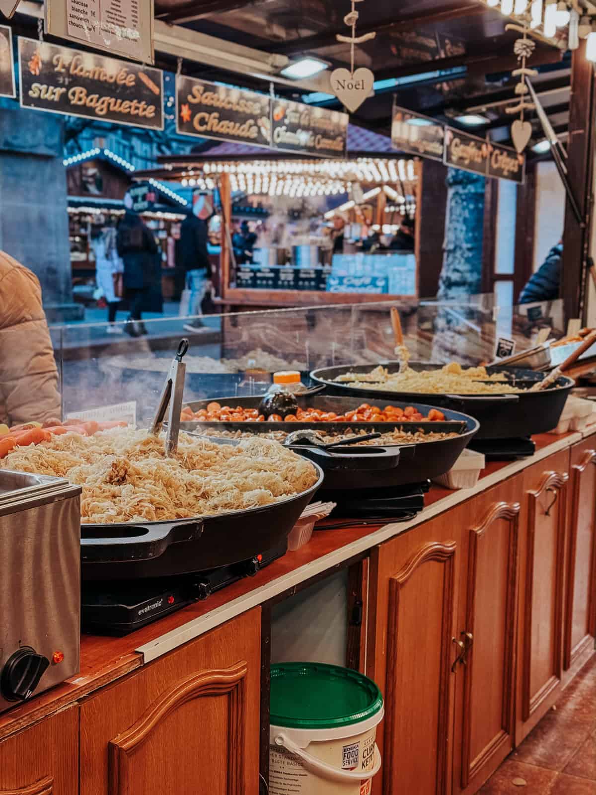 A Christmas market food stall in Kaysersberg offering steaming dishes of sauerkraut and sausages, with festive signs and a merry atmosphere reflected in the shiny countertops.