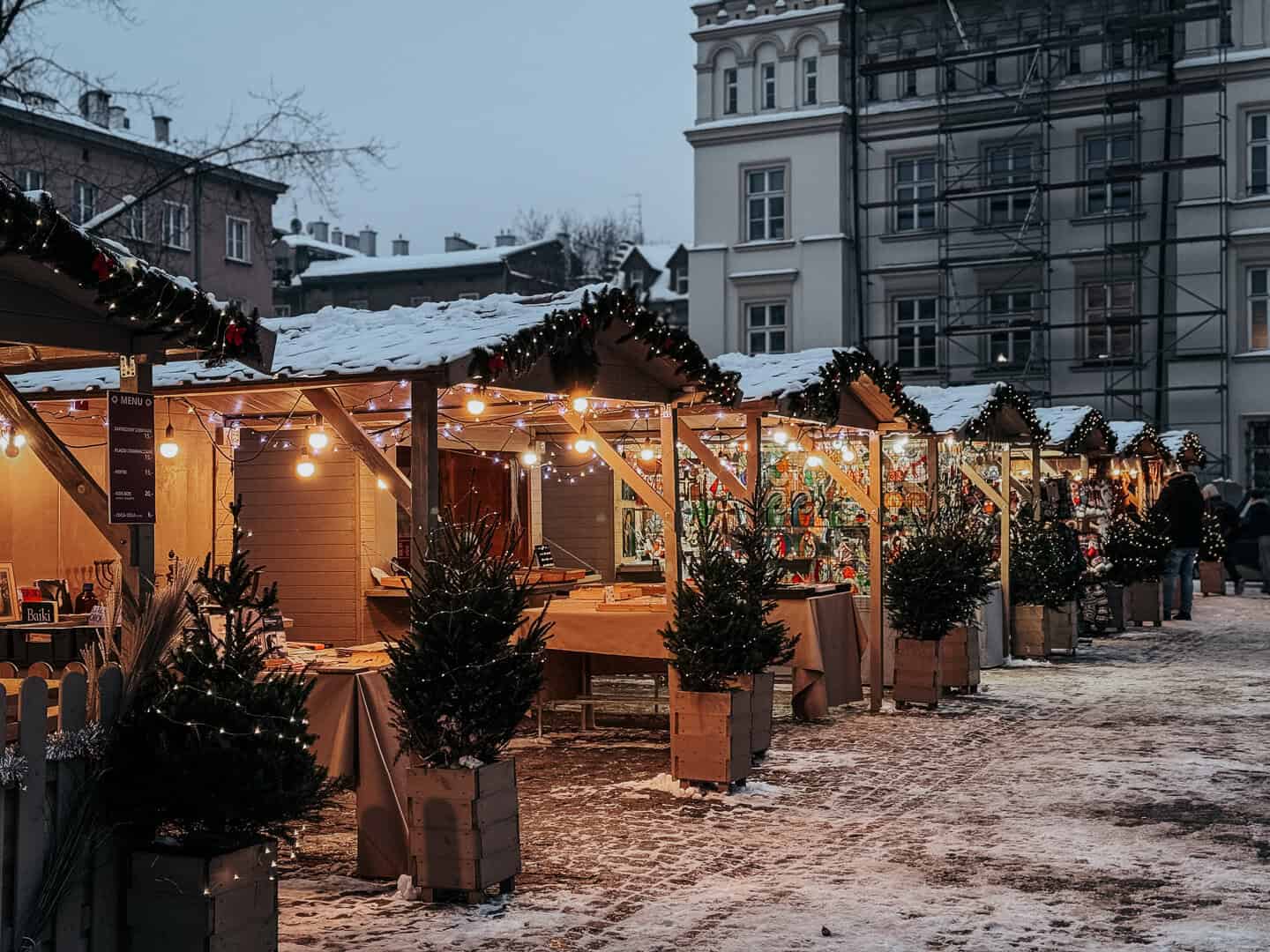 Twilight over a snow-dusted Christmas market in Krakow with a row of wooden stalls decorated with lights and wreaths