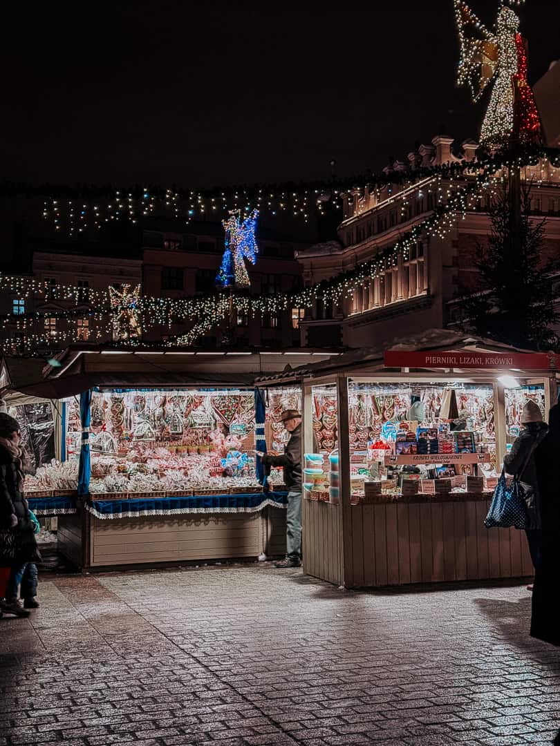 Illuminated Christmas market stalls at night with festive decorations and a large lit angel figure in the backdrop in Krakow.