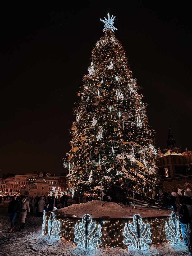 A towering Christmas tree adorned with lights, ornaments, and a bright star topper, surrounded by people in a Krakow square