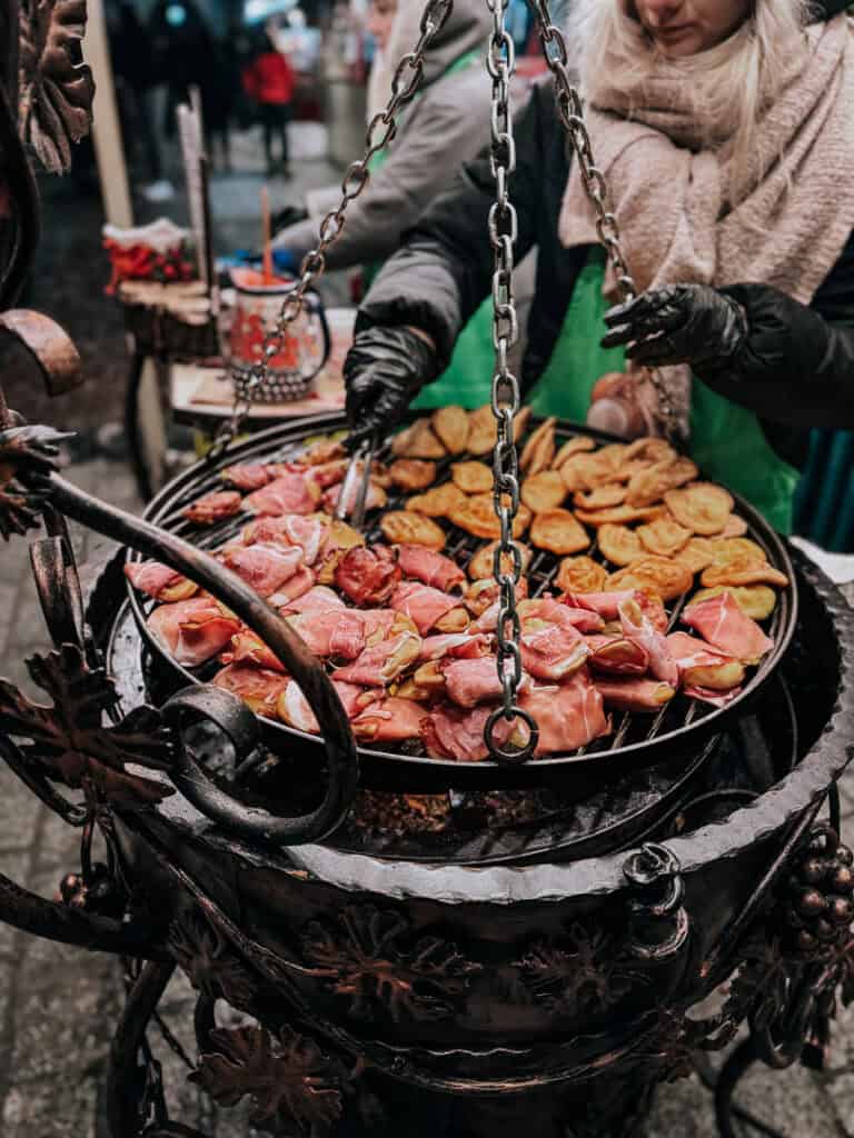Close-up of a traditional open grill cooking various meats and potatoes, tended by vendors at a Christmas market in Krakow