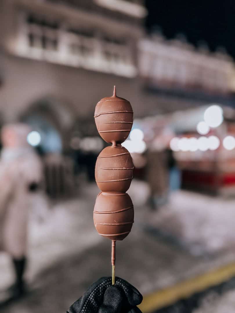 A hand holding a skewer of four chocolate-covered marshmallows against a blurred city night background
