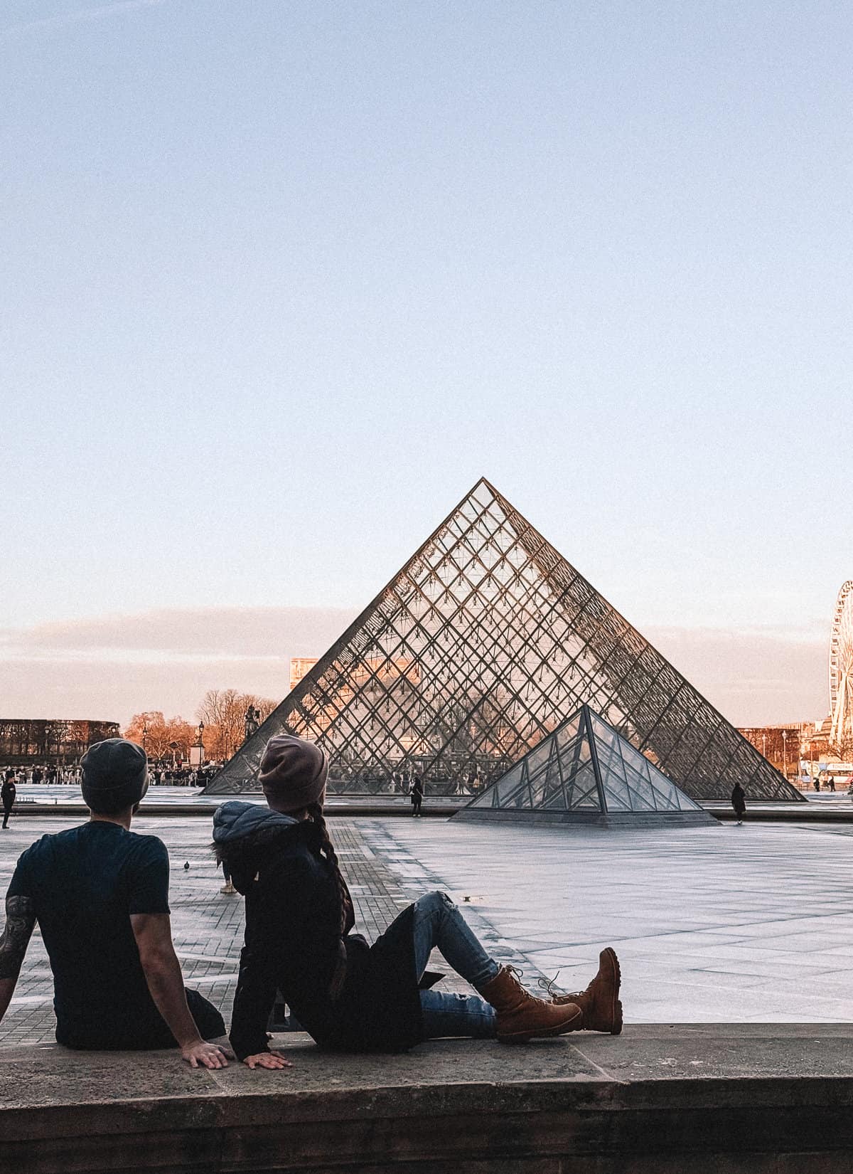 A couple sits back-to-back on the edge of the fountain at the Louvre, with the museum's famed glass pyramid and a clear blue sky at dusk.