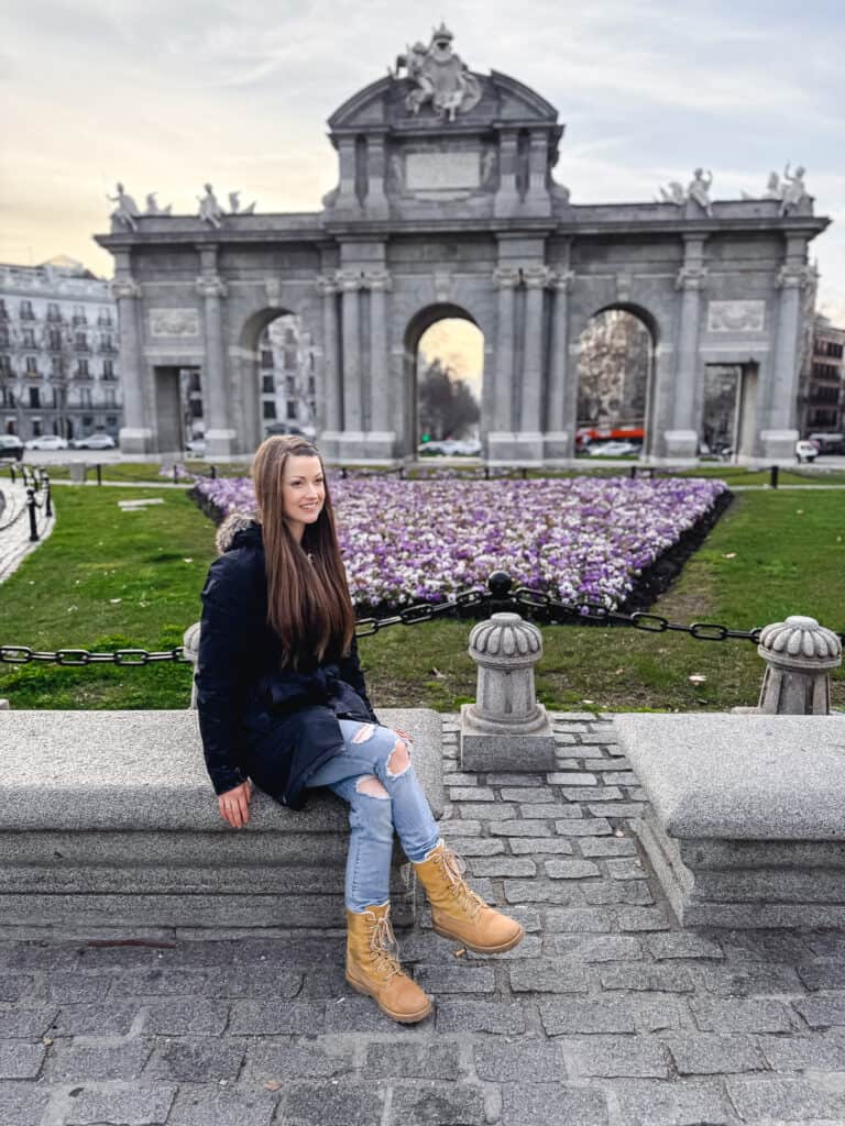 A woman sits in front of puerta de alcala in madrid surrounded by purple flowers