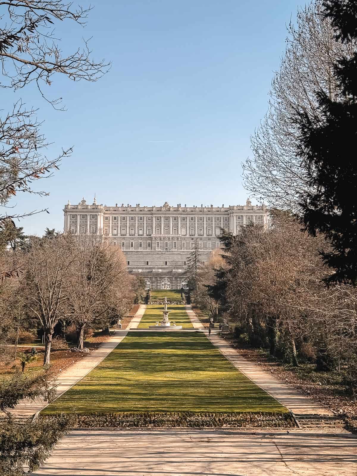 View of the Royal Palace of Madrid from the manicured green gardens of Campo del Moro, with a clear blue sky above, showcasing the expansive size and neoclassical architecture of the historic Spanish residence