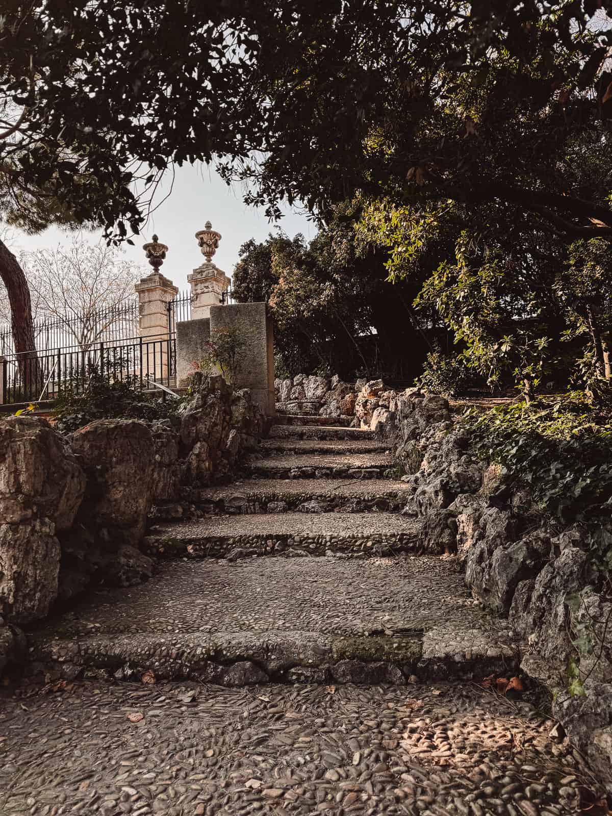 A serene and rustic stone staircase leading through a leafy garden with mature trees, glimpses of ornate fencing, and classical stone finials, suggesting a quiet retreat within a Madrid park.