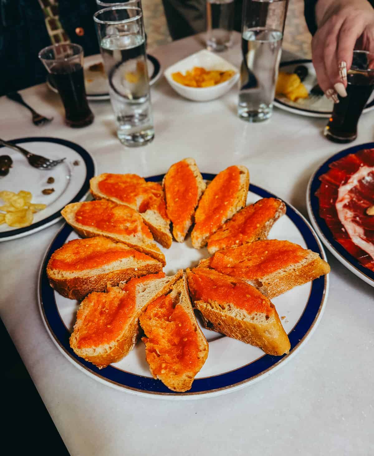 A plate of toasted bread topped with a rich, orange tomato spread, served on a white and blue rimmed plate beside a tray of sliced ham and other tapas, indicating a typical Spanish meal.
