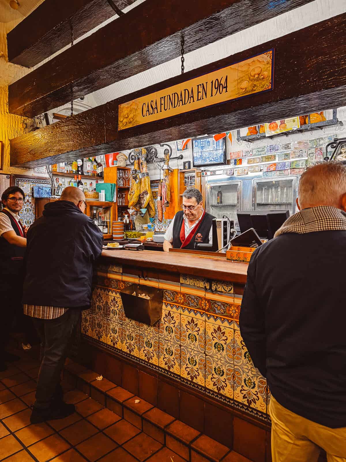 Inside a cozy Spanish mesón, patrons engage with a bartender beneath a wooden sign dating the establishment to 1964, surrounded by ornate tilework and various hanging meats and tapas.