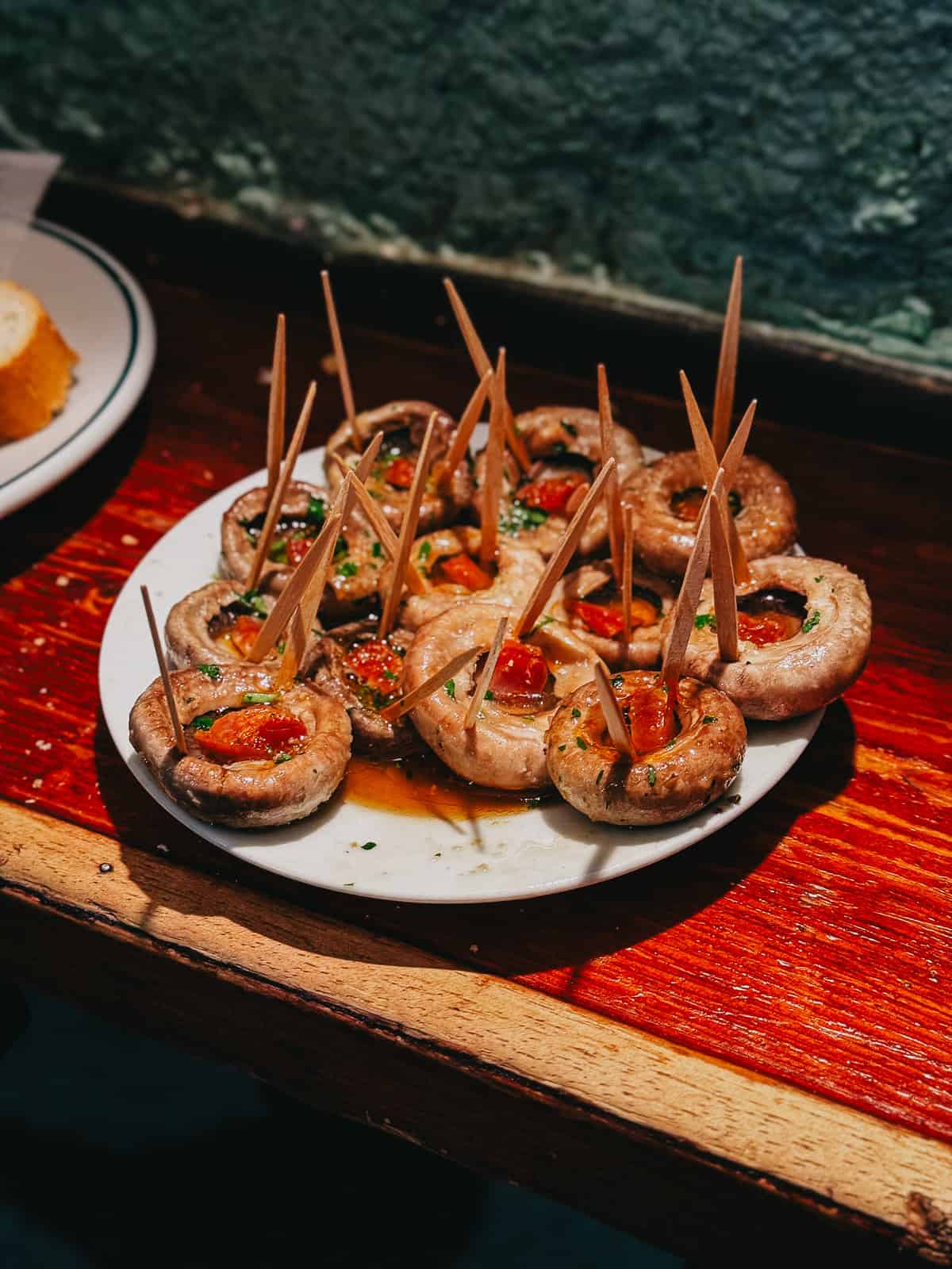 A close-up of grilled mushrooms topped with herbs and tomato on a white plate with toothpicks, served as tapas on a wooden bar counter, embodying a traditional Spanish appetizer.