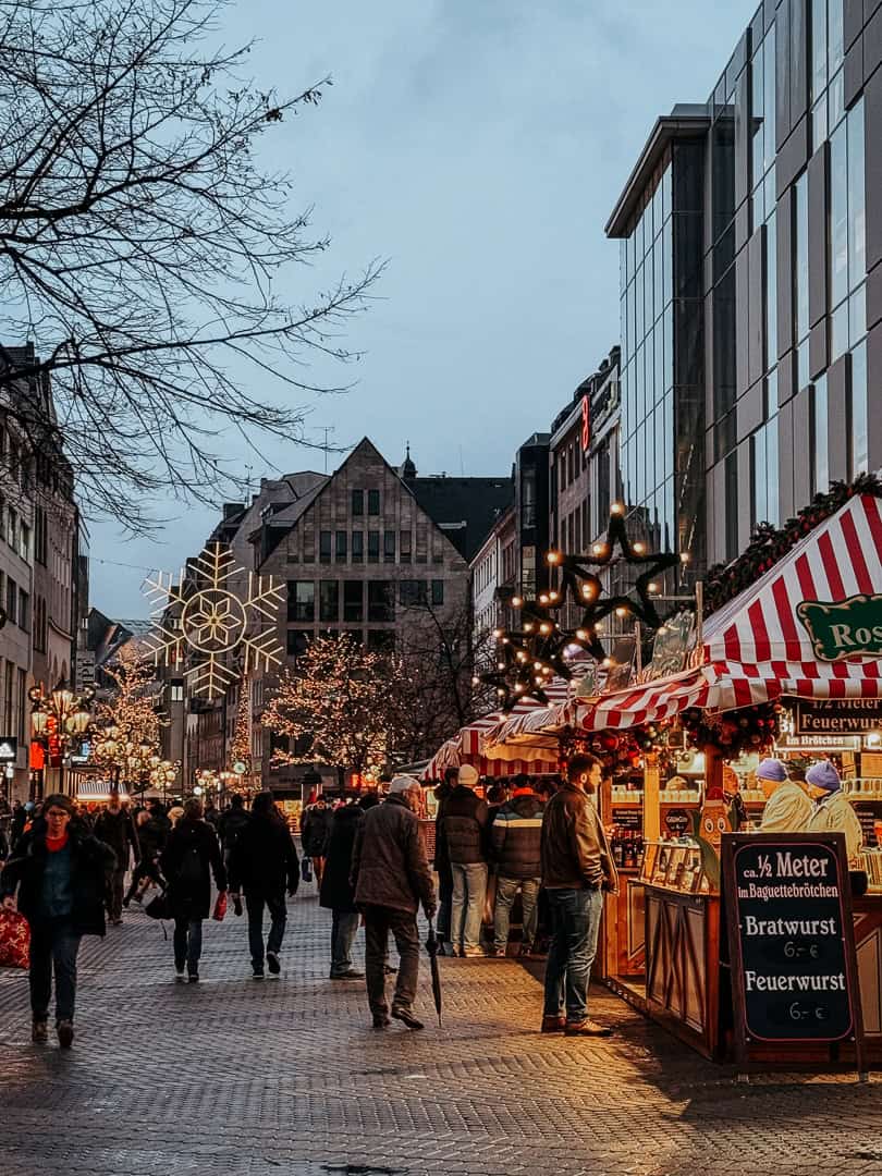A bustling Christmas market street in Leipzig with people, festive lights, and a stall selling 'Bratwurst' and 'Feuerwurst' in the evening.