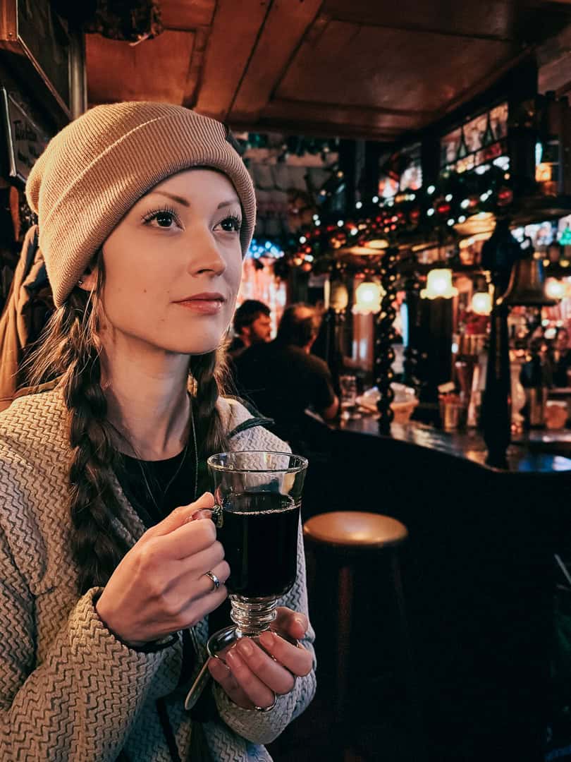 A woman wearing a beanie holds a glass of mulled wine, with a cozy, decorated pub interior in the background
