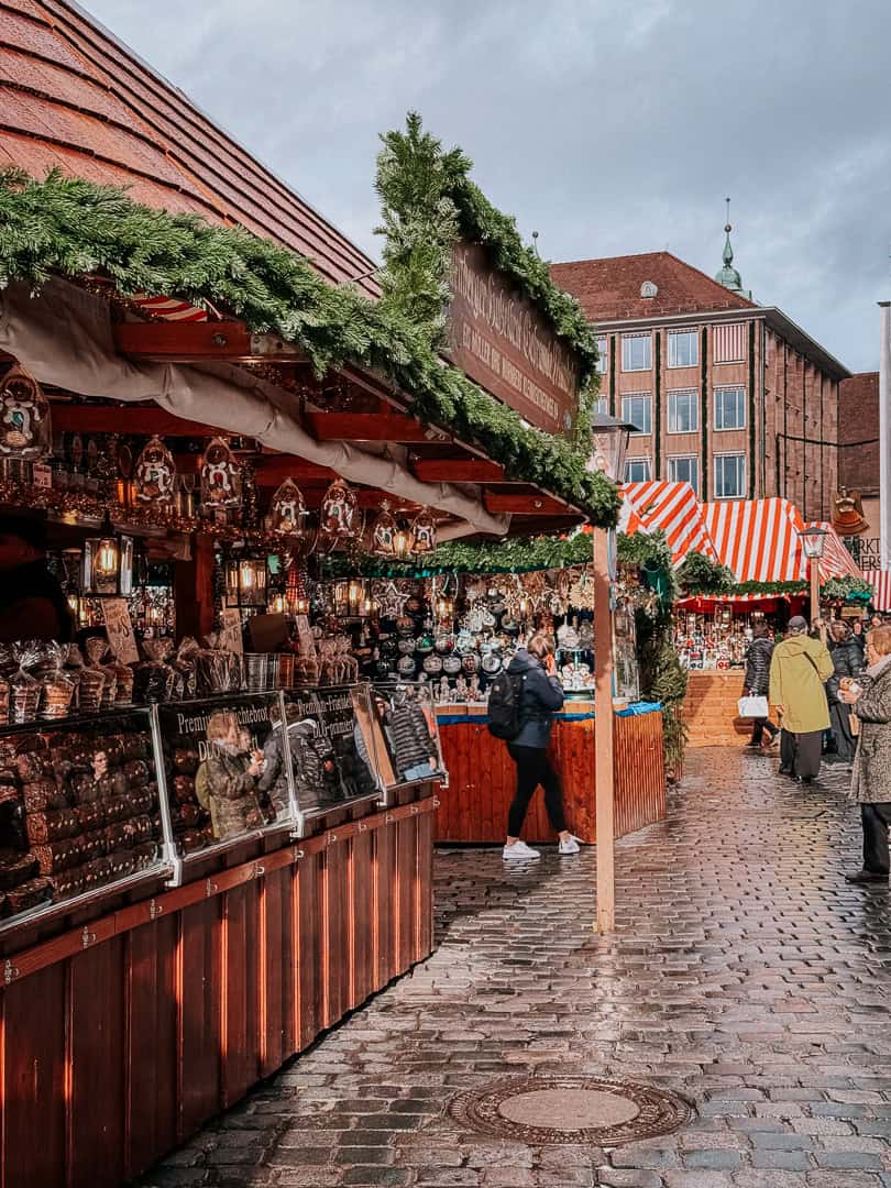 A bustling Christmas market in Nuremberg with traditional decorations and a variety of gingerbread on display under a sign for 'Lebkuchen Schmidt'.