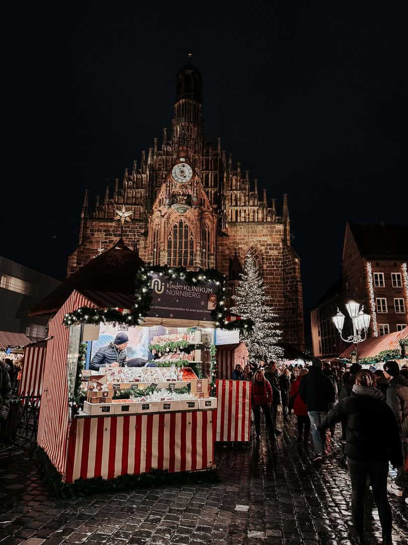 A festive Christmas market stall at night selling seasonal treats with the illuminated Gothic spire of the Frauenkirche in Nuremberg as a backdrop.