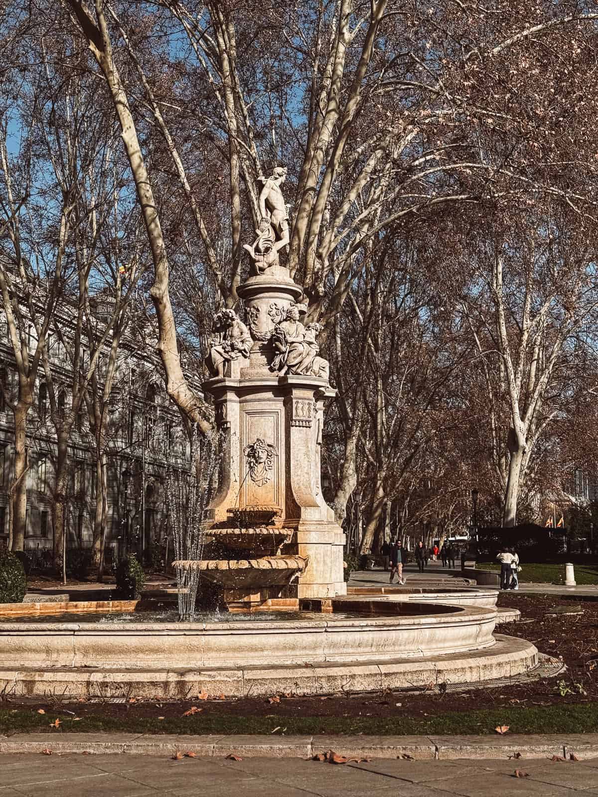 An ornate stone fountain adorned with sculptures, centered among bare-branched trees under a blue sky, with water gently cascading down its tiers.
