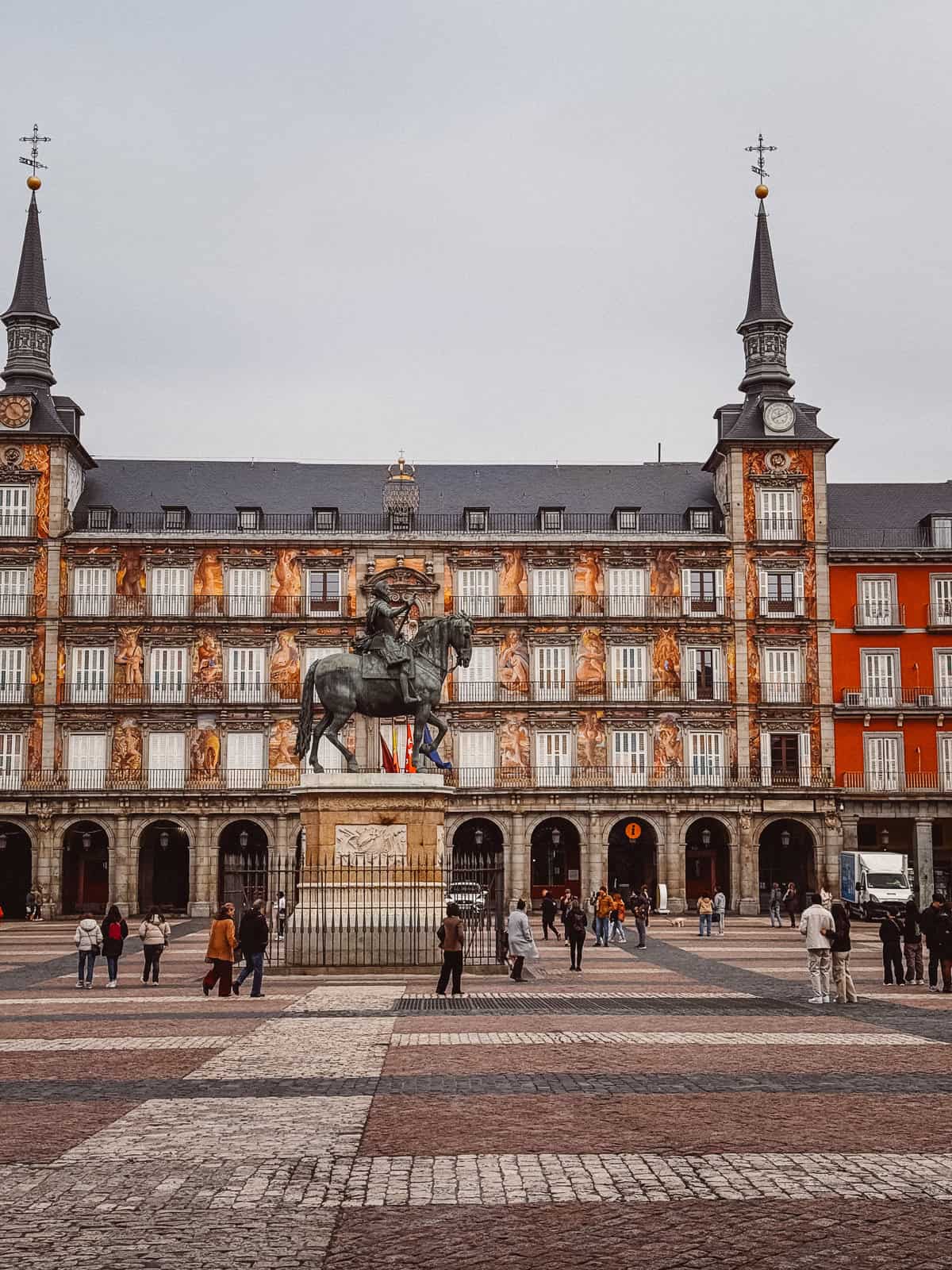 Visitors gather in the grand Plaza Mayor of Madrid, featuring the iconic equestrian statue of King Philip III set against the backdrop of the plaza's traditional Spanish architecture with ornate facades and spired towers under a cloudy sky