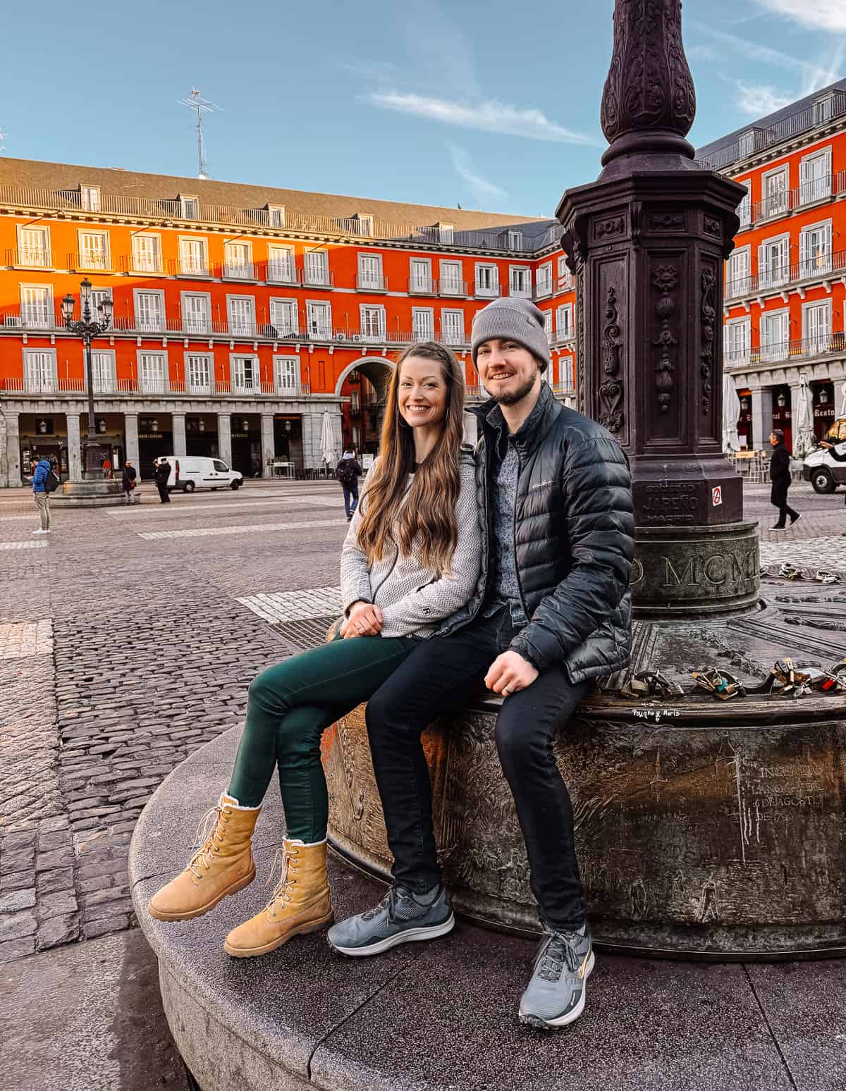 A joyful couple sitting on the edge of a fountain at Plaza Mayor in Madrid, the iconic red façades of the surrounding buildings framing their relaxed, contented pose.