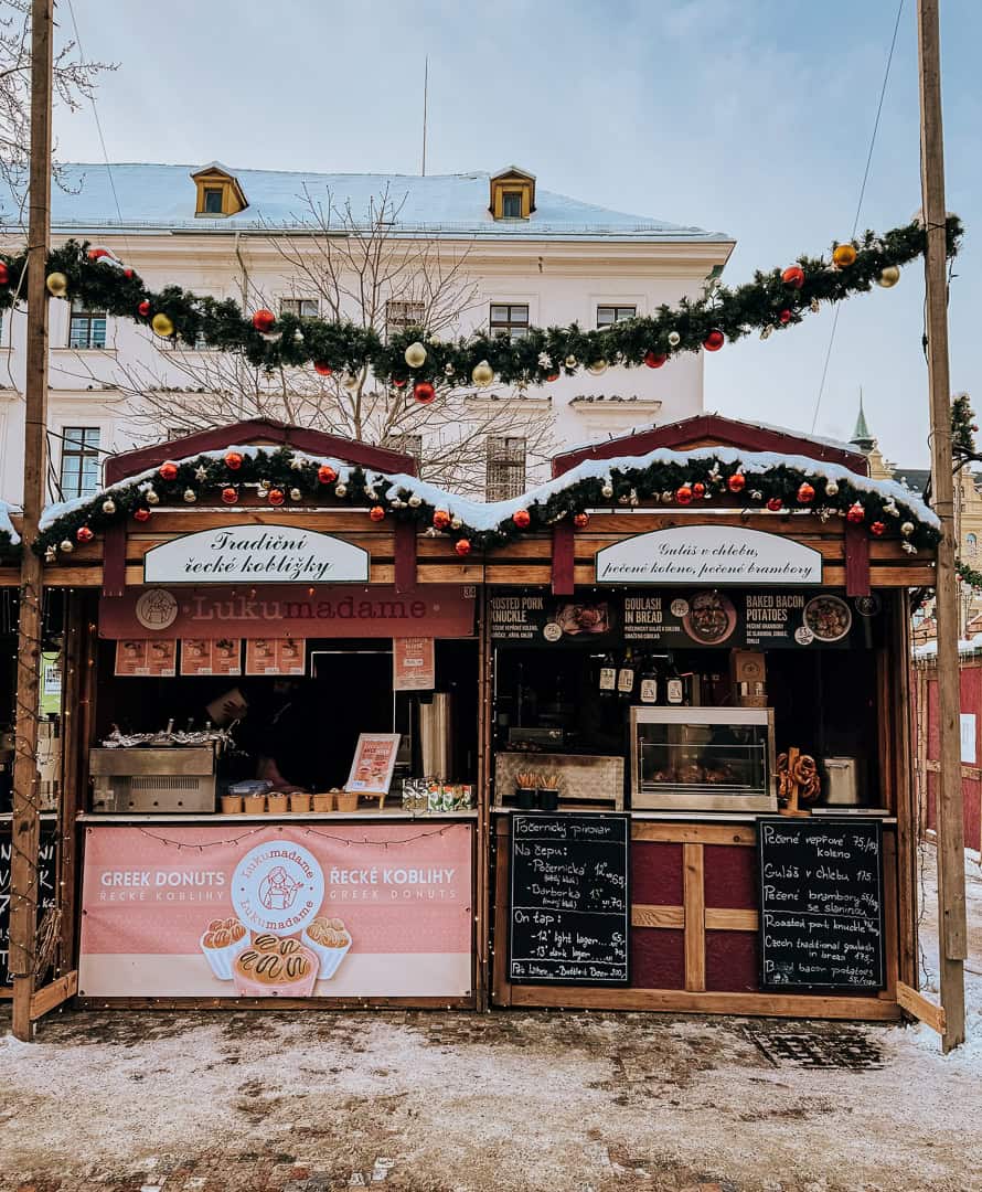 two quaint christmas market booths with wooden fronts and garlands stand side by side at a market in Prague. One sells mulled wine and one sells sweets
