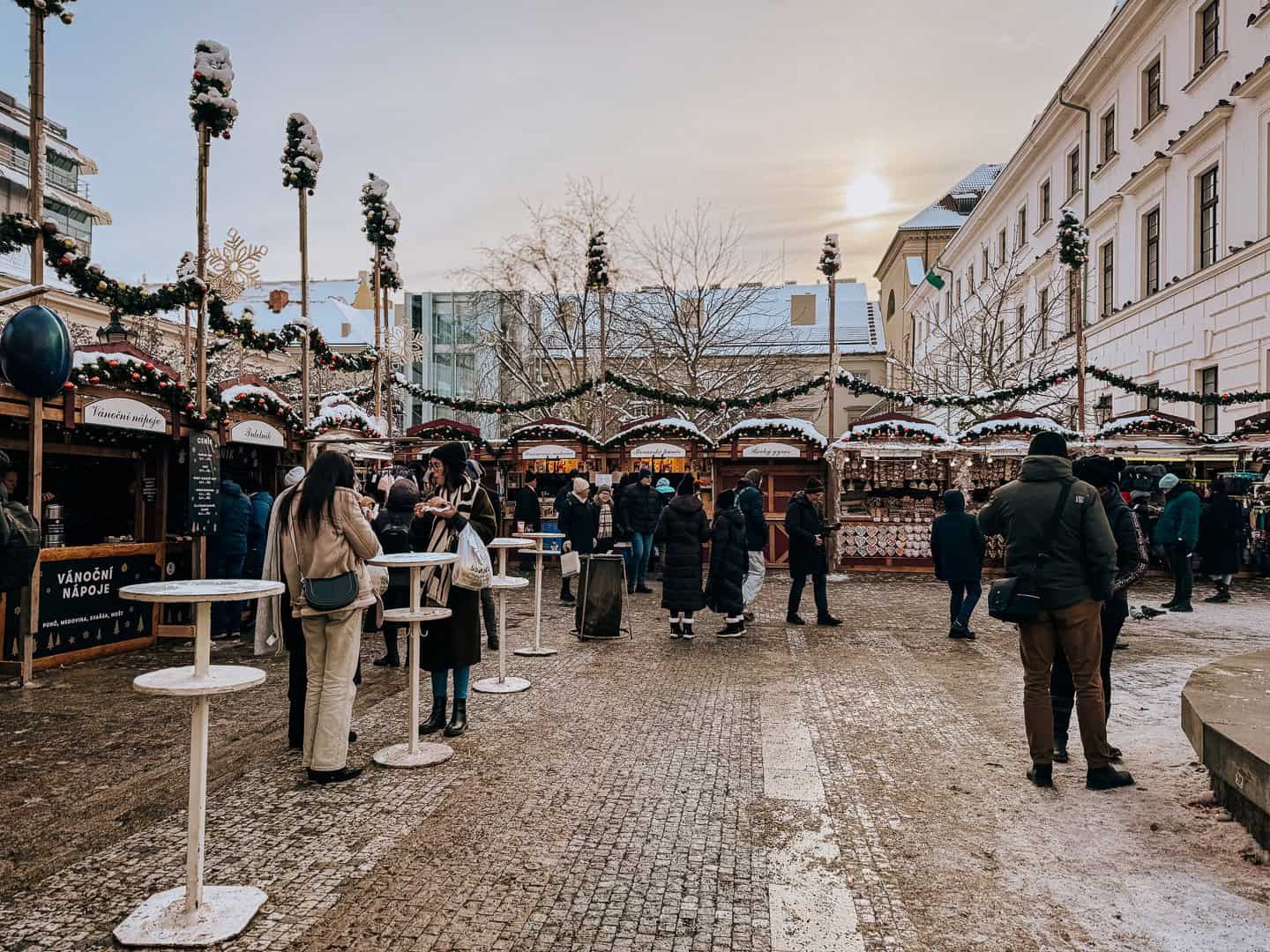 A bustling Christmas market during daylight with shoppers walking among festively decorated wooden stalls under a soft winter sun.