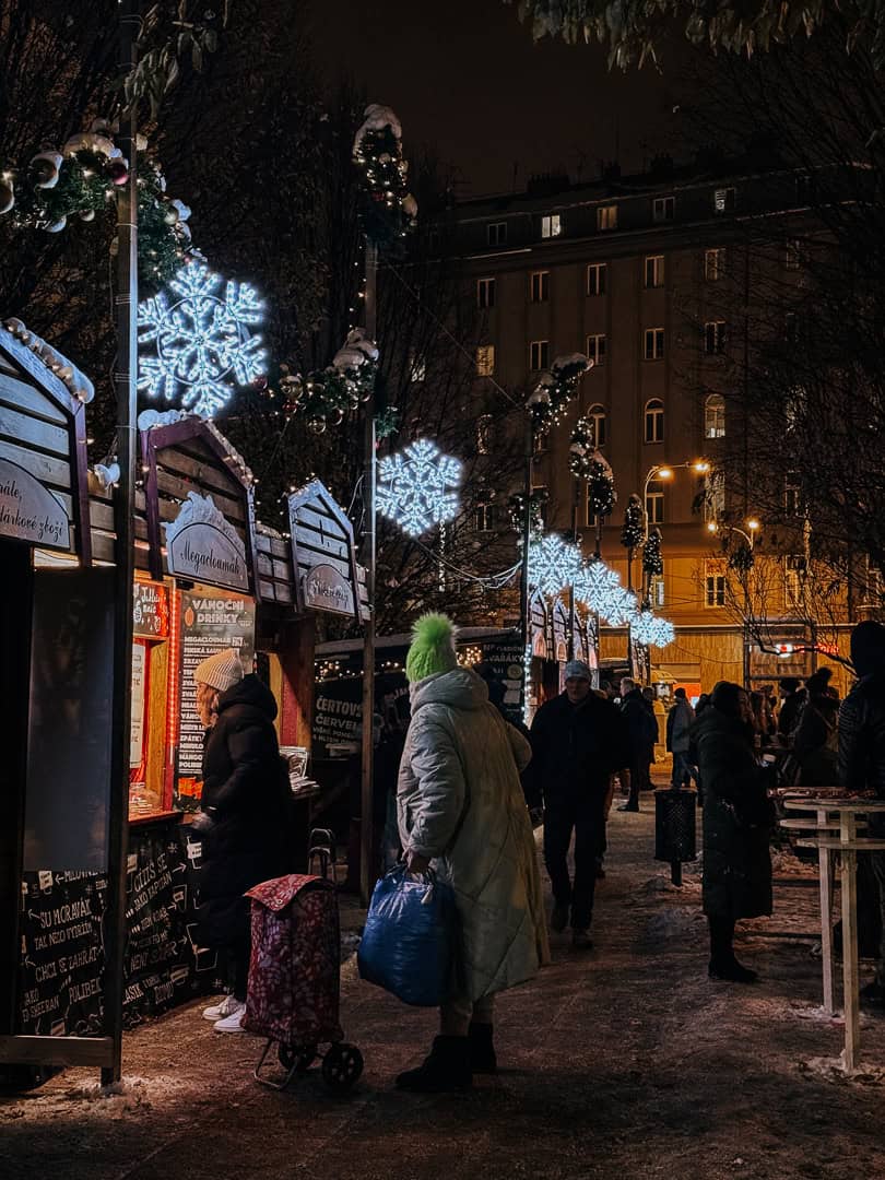 Shoppers in winter attire browsing through a Christmas market at night, illuminated by ornate light decorations and a glow from the festive wooden huts.
