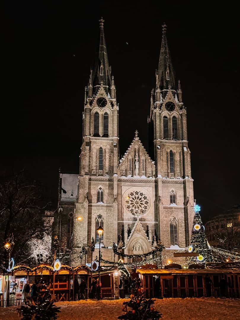 A majestic Gothic church towers over a lively Christmas market, its spires lit against the night sky, with holiday shoppers milling about the booths below.
