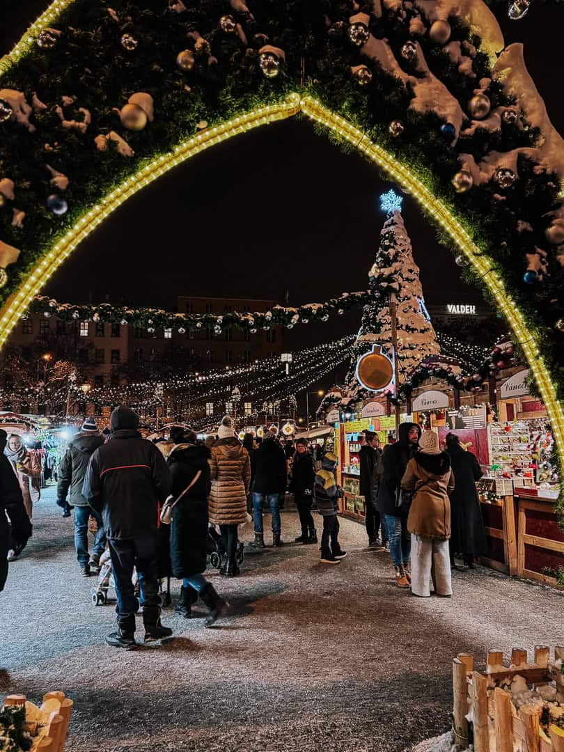 Visitors walking under a grand illuminated archway at a Christmas market, with a towering Christmas tree and festive booths in the background.