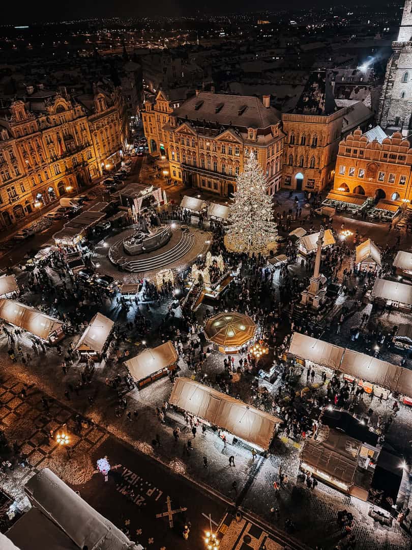 A high-angle night shot of Prague's Christmas market bustling with people around a central Christmas tree, adjacent to ornate historical buildings.