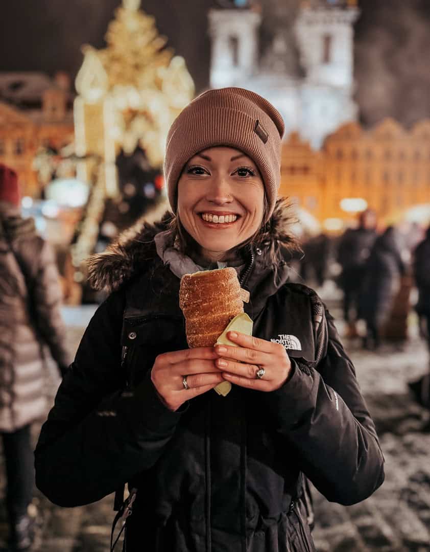 A smiling woman in a winter hat holds a traditional Czech trdelník pastry, with a festive Christmas market and a glowing tree in the background.