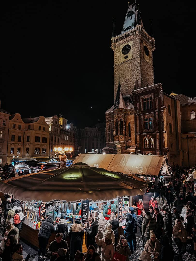 A busy evening scene at the Christmas market in Prague's Old Town Square, with the Gothic tower of the Old Town Hall rising above the crowd.