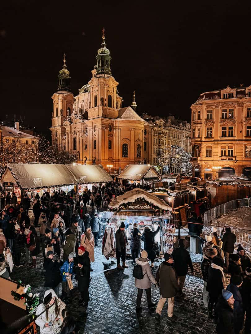 Snow-covered roofs of Christmas market stalls with crowds of visitors enjoying the festive atmosphere near the Baroque Saint Nicholas Church in Prague