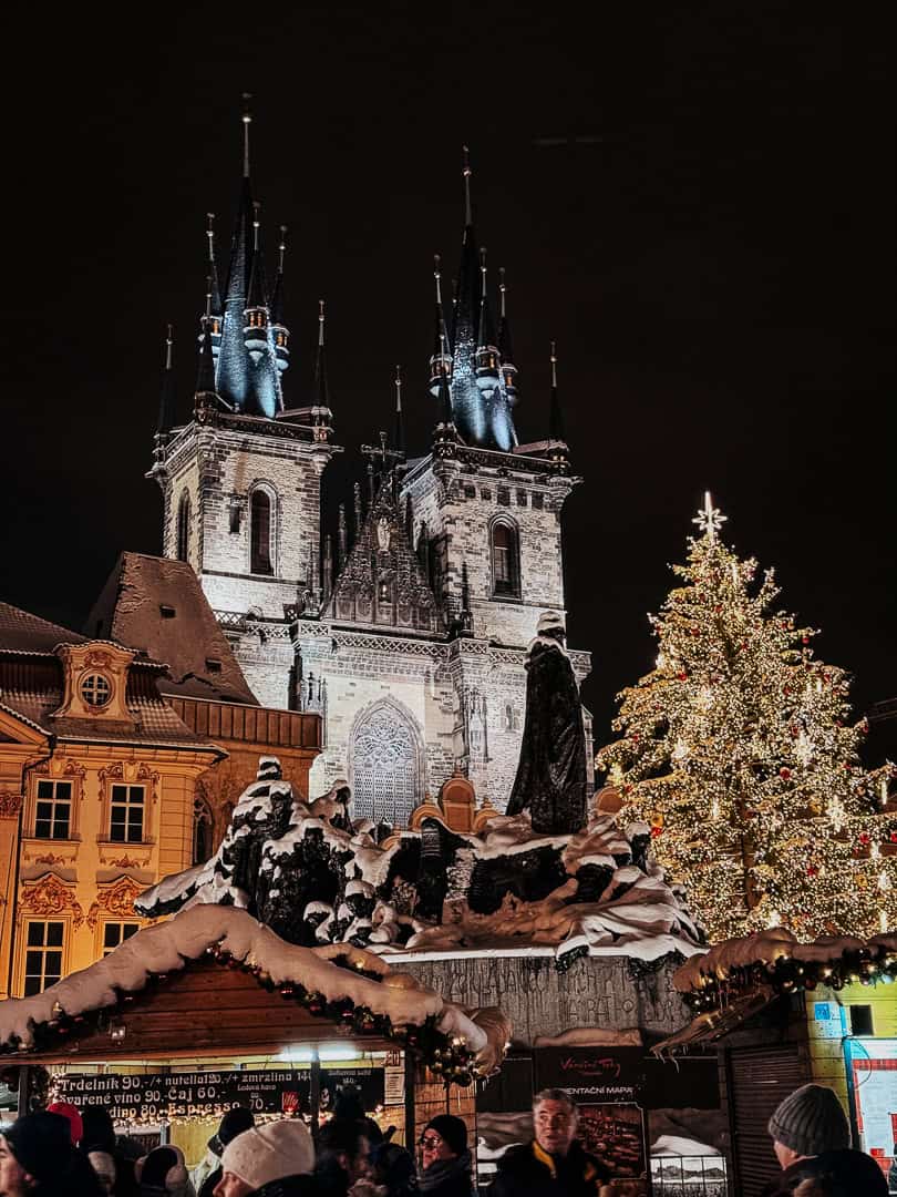 The iconic Church of Our Lady before Týn in Prague, dramatically lit against the night sky, overlooks the bustling Christmas market below