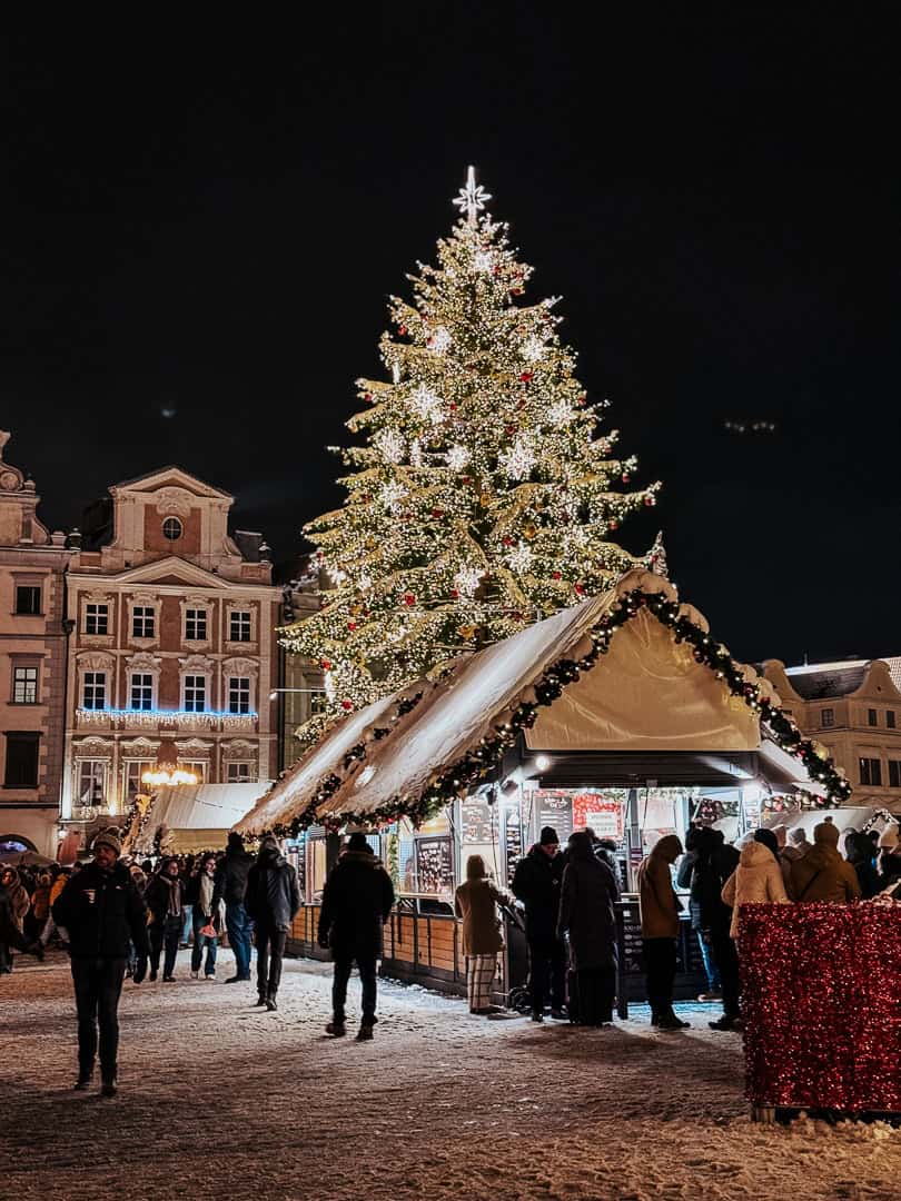 A brightly lit Christmas tree stands tall in the evening at a snow-covered Prague Christmas market, with festive stalls and a crowd of shoppers around.