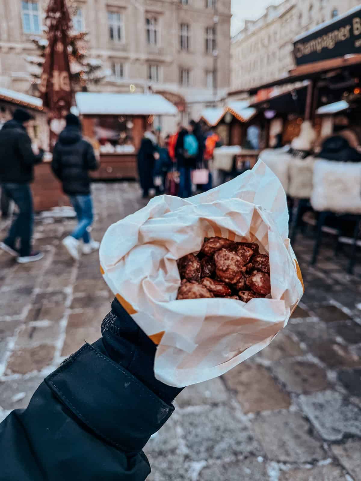 A first-person view of a paper cone filled with caramelized roasted almonds held in a gloved hand, with a blurred Christmas market scene in the background
