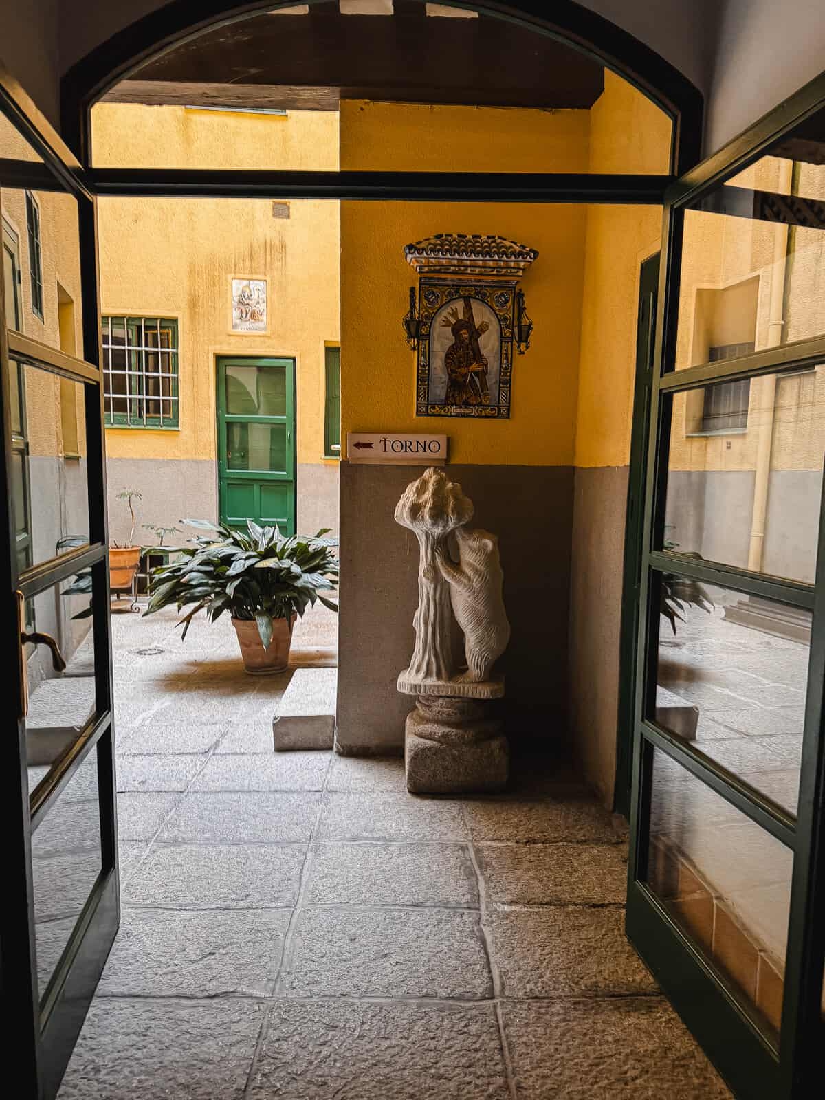 Interior courtyard view through an open doorway, showcasing a statue of a robed figure next to a 'TORNO' sign, with a decorative tile depicting a saint on the yellow wall.