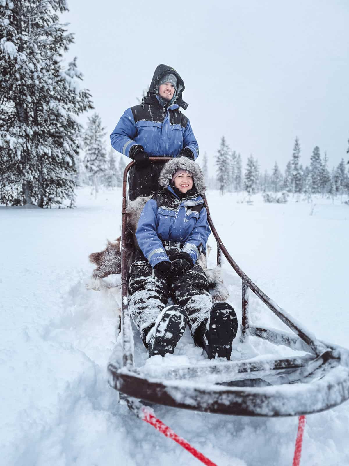 A joyful couple experiences dog sledding, with one steering the sled and the other seated comfortably, as they travel through a snowy winter wonderland.