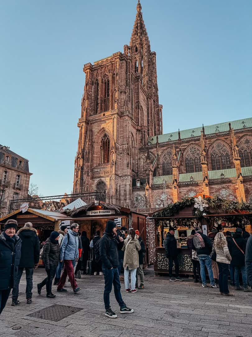 Visitors stroll past a magnificent Gothic cathedral towering over a Christmas market, with vendors' stalls offering seasonal delights under a clear blue sky.