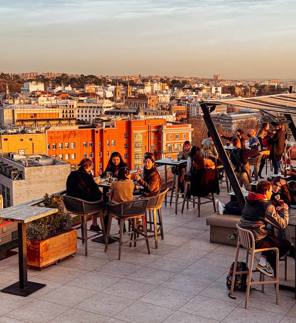 Patrons enjoy a rooftop dining experience, basking in the golden light of the setting sun with a panoramic view of the city's horizon as their backdrop.
