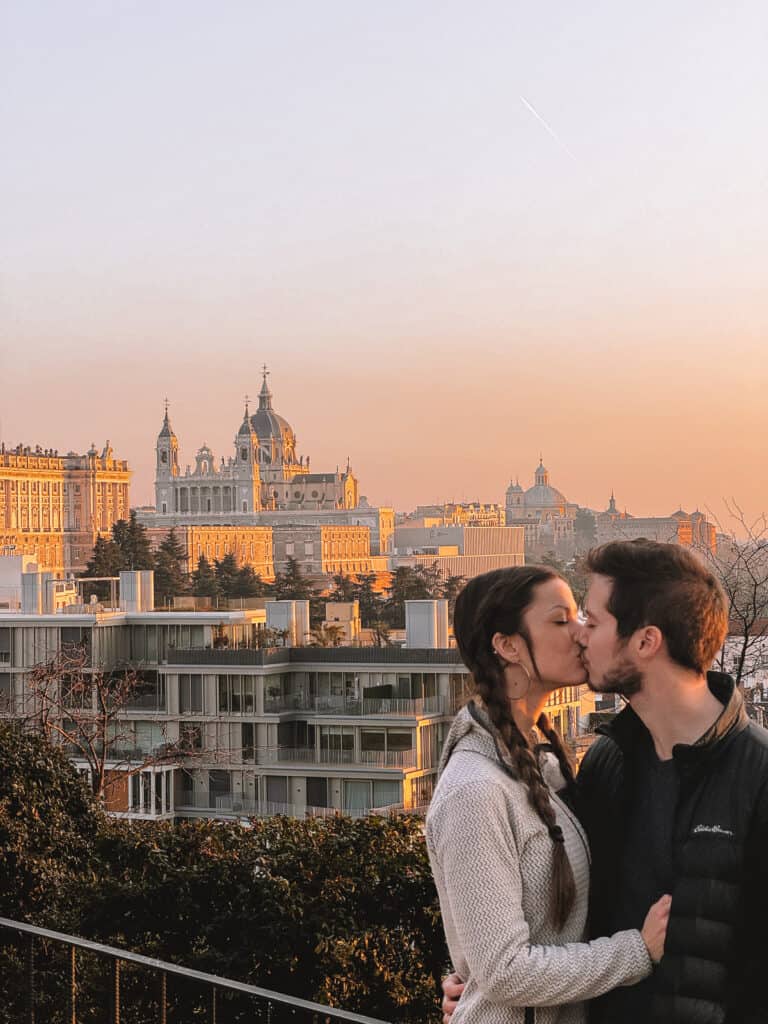 A couple sharing a kiss with the Madrid skyline in the background during sunset, highlighting a romantic moment against a scenic urban backdrop.