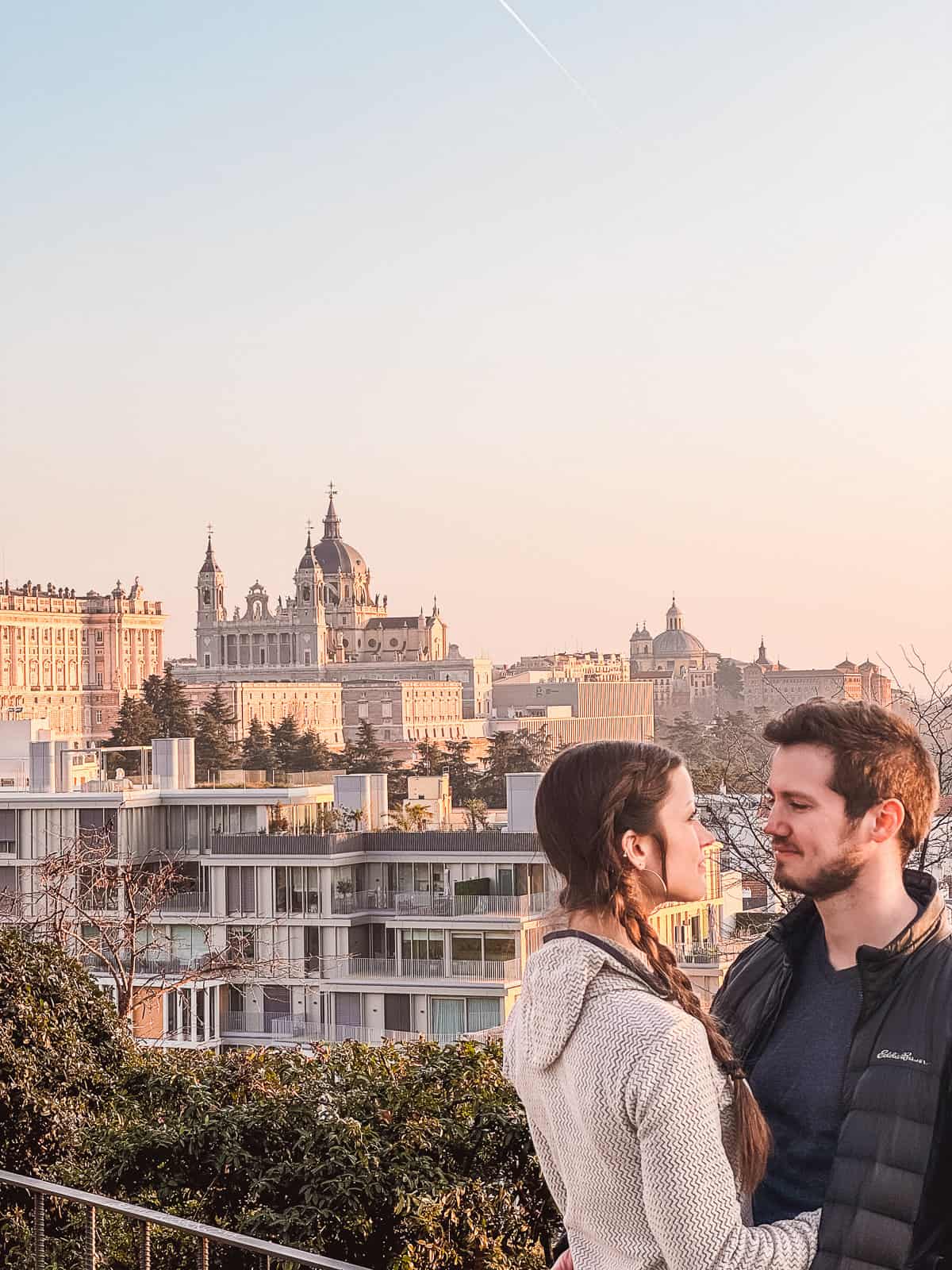 A couple gazing into each other's eyes in the foreground with the historical Templo de Debod and Madrid's cityscape bathed in the warm glow of sunset in the background.