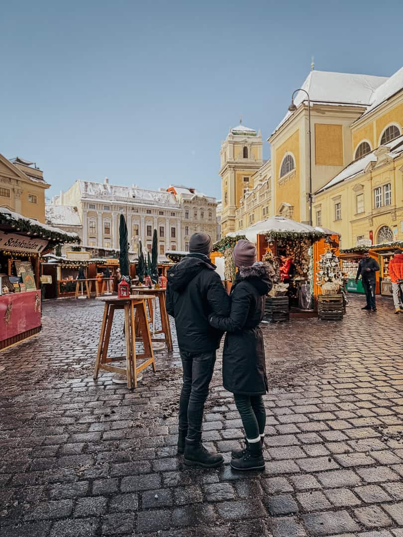A couple in winter clothing observes a Christmas market in Vienna, with historical yellow buildings framing the scene under a bright blue sky.